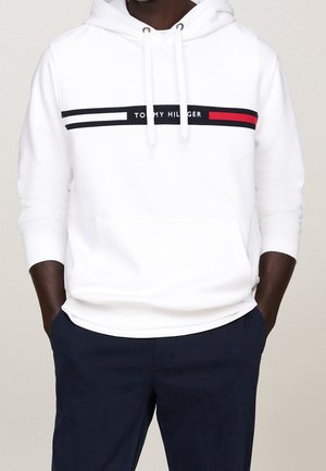 Man draagt een witte Tommy Hilfiger-hoodie met een logostreep over de borst en donkerblauwe broek, handen in de zakken.