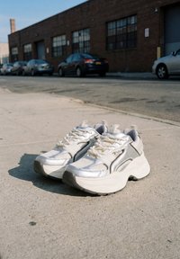 Baskets épaisses argentées et blanches posées sur un trottoir urbain ensoleillé avec des voitures garées et un bâtiment en briques en arrière-plan.