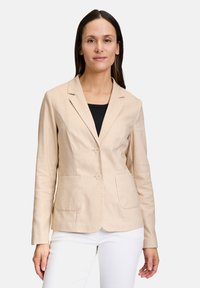 Beżowa marynarka wykonana z lnianego materiału, dopasowana, z dwiema przednimi kieszeniami i dwoma guzikami z przodu. Nosi się ją na czarnym topie.