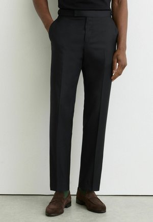 Muž oblečený v čiernych na mieru šitých nohaviciach, zelených ponožkách a hnedých semišových mokasínach stojaci opretý o bielu stenu s uvoľnenými rukami.