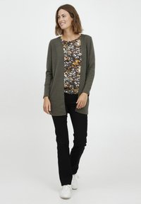 Olivgrön lång cardigan över en blommig topp i varma nyanser, kombinerad med svarta byxor och vita sneakers, med en avslappnad passform.