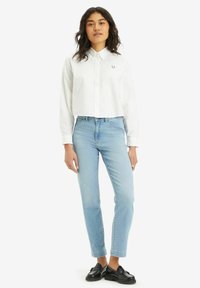 Camicia bianca con colletto e dettagli ricamati, abbinata a jeans chiari a sigaretta e mocassini neri. Sfondo semplice.