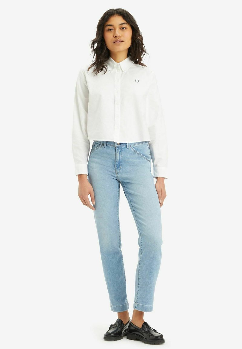 Weißes Hemd mit Kragen und besticktem Detail, kombiniert mit hellblauen schmal geschnittenen Jeans und schwarzen Slippern. Schlichter Hintergrund.
