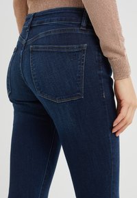 Mörkblå denimjeans med en figurnära passform, med två bakfickor och synliga sömnadsdetaljer. Tyget har en slät textur.