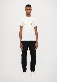 Balta marškinėliai su auksiniu "Versace Jeans Couture" logotipu, derinami su juodomis kelnėmis ir baltomis sportbačiais, demonstruojami ant modelio prieš vientisą foną.