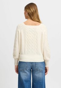 Kremni pleteni pulover z kabelskimi vzorci, širokimi rokavi in dizajnom s krojenim hrbtom, v kombinaciji z modrimi širokimi jeans hlačami.