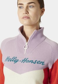 Sweter z wysokim kołnierzem, w kolorze lawendowym, białym i czerwonym w stylu color-block. Posiada zapięcie na zamek oraz logo "Helly Hansen" w kolorze turkusowym.