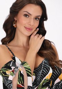 Kvinde med langt bølget brunt hår iført store guldøreringe og en farverig mønstret off-shoulder top, der rører ved sit ansigt med et let smil.