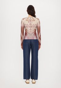 Langärmliges, eng anliegendes Hemd mit komplizierten, multicolor Grafiken. Kombiniert mit weit geschnittenen Jeanshosen und weißen Schuhen. Neutraler Hintergrund.