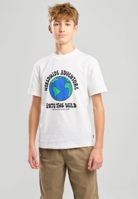 Camiseta de algodón blanca con un gráfico colorido del globo rodeado de texto, con mangas cortas y un cuello redondo. Se lleva con pantalones beige.