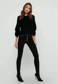 Suéter preto canelado com mangas balão, combinado com calças skinny pretas de cintura alta com cinco botões e botas de salto preto até o tornozelo.