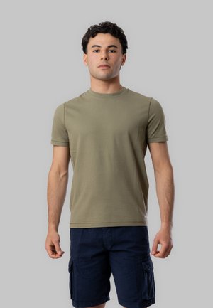 Giovane uomo con capelli corti e scuri, che indossa una maglietta verde oliva e pantaloncini cargo blu navy, in piedi davanti a uno sfondo grigio uniforme.