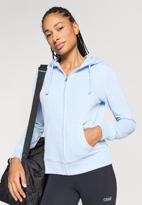 Σιέλ φούτερ με φερμουάρ, κορδόνια και μπροστινές τσέπες, κατασκευασμένο από απαλό ύφασμα. Φοριέται με μαύρα αθλητικά κολάν και κρατάει μια μαύρη τσάντα duffel.