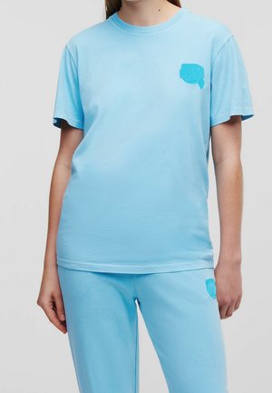 Osoba nosząca jasnoniebieski t-shirt z krótkim rękawem i dopasowane spodnie, oba z małymi niebieskimi haftowanymi naszywkami w pobliżu klatki piersiowej i uda.