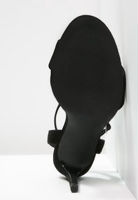 Musta korkokenkä, jossa on kuvioitu pohja, avoin takapuoli ja ohuet hihnat. Nukka materiaali lisää pehmeän tekstuurin suunnitteluun.