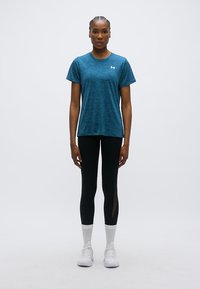 Αθλητική μπλούζα σε τιρκουάζ χρώμα από ελαφρύ, υδρόφιλο ύφασμα, συνδυασμένη με μαύρα κολάν που έχουν πλέγμα στο πλάι και λευκά αθλητικά παπούτσια.