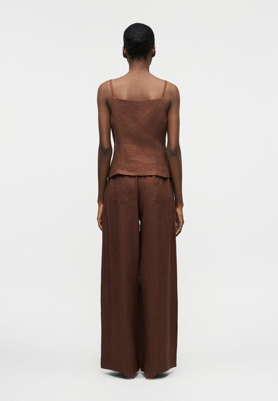 Γυναίκα με σκούρο δέρμα και μαλλιά δεμένα πίσω, φορώντας αμάνικη καφέ μπλούζα και παντελόνια με φαρδιά πόδια, στέκεται με την πλάτη στραμμένη προς ένα απλό φόντο.