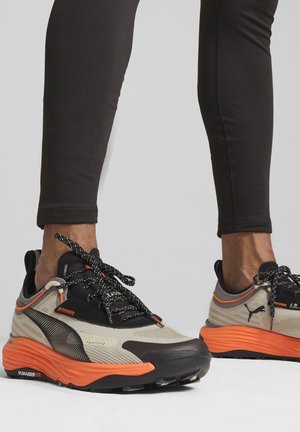 Sportssko med en beige mesh-overdel, sorte detaljer og en orange gummisål. Teksturerede snørebånd og logo-detalje på siden.