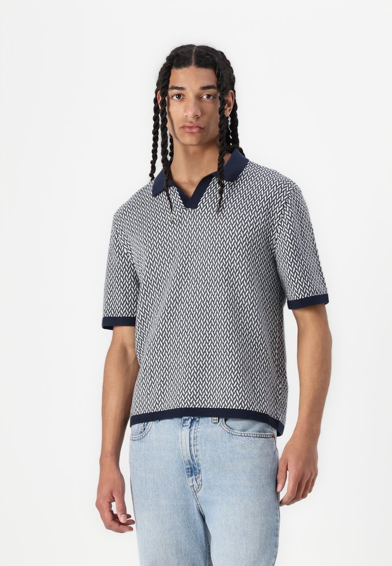 Polo de punto de manga corta en patrón zigzag azul marino y blanco, con cuello y puños en contraste azul marino. Textura suave, ajuste relajado.