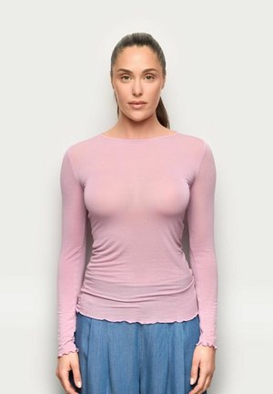 Kobieta z brązowymi włosami związanymi do tyłu, ubrana w jasnoróżową bluzkę z długim rękawem i niebieskie spodnie, stojąca na jednolitym białym tle.