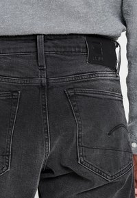 Pantalones vaqueros de mezclilla gris oscuro con un acabado texturizado, que cuentan con una parche de cuero negro en la cintura trasera y dos bolsillos traseros con detalles de costura.