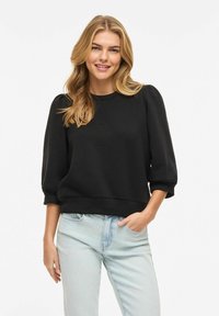 Femme souriante aux longs cheveux blonds portant un pull noir à manches bouffantes et un jean bleu clair, debout avec une main dans la poche.