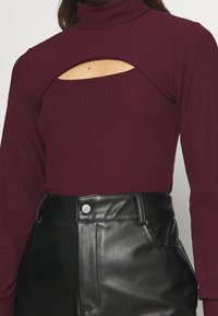 Burgundy ribbad långärmad topp med en hög krage och utskärning vid bröstet, i kombination med högmidjade svarta imitationläderbyxor.