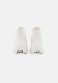 Białe sneakersy typu high-top z gumową podeszwą, z teksturowanym materiałem, okrągłymi akcentami logo oraz falistym wykończeniem na kołnierzu.