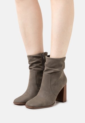 Bottes chevilles en suédine grise avec dessus bouffant, fermeture éclair latérale, bouts pointus et talons en bois empilés. Texture lisse avec un léger éclat.