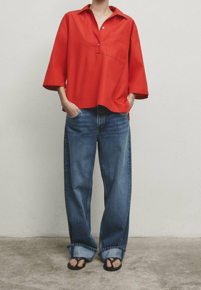 Camicia rossa di cotone con apertura a bottoni e maniche corte, abbinata a jeans in denim blu a gamba larga e infradito neri.