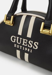 Sort læderhåndtaske med beige dobbelte striber, guld hardware og "GUESS EST 1981" logo foran.