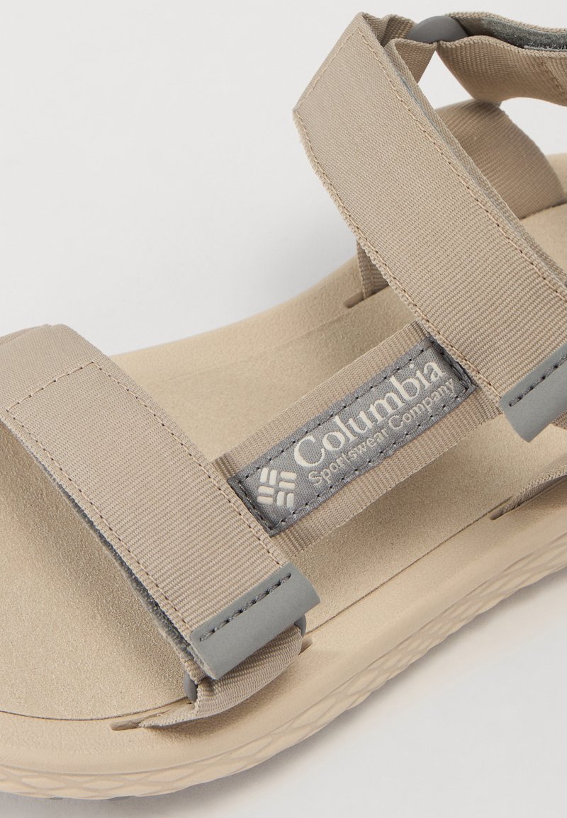 Sandalia beige con cintas grises, con un upper de textil, cierres de velcro ajustables y una plantilla de espuma acolchada. Marca Columbia visible.