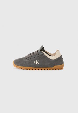 Zapatilla de ante gris con suela de goma. Presenta una pestaña en el talón negra, un acento beige en la parte trasera y un logo dorado en el lateral. Diseño con cordones.
