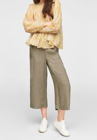 Blusa gialla a fiori con pieghe, abbinata a pantaloni larghi in lino verde oliva e sneakers bianche.