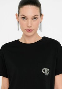 T-shirt nera con scollatura rotonda e tasca decorata con un logo impreziosito da cristalli. Il modello indossa orecchini a catena lunghi e argentati.
