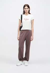 Jovem mulher com cabelo escuro veste uma t-shirt branca da Nike, calças de moletom cor malva, ténis brancos e uma mala de ombro preta, de pé contra um fundo simples.