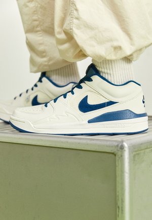 Białe i niebieskie buty sportowe Nike noszone z beżowymi spodniami i białymi skarpetkami, stojące na zielonej metalowej platformie.