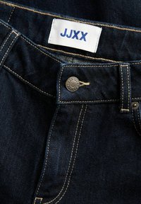 Tmavé džíny z denimu se zlatým prošíváním, s stříbrným knoflíkem a bílou etiketu s modrým textem "JJXX" poblíž pasu.