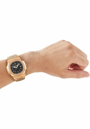 Mandligt håndled iført et guldfarvet metalarmbåndsur med sort urskive og hvide tal, sat mod en hvid baggrund.