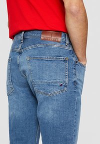 Jeans de mezclilla azul claro con una textura suave. Cuenta con un parche de cuero marrón y un pequeño acento de logotipo cerca del bolsillo trasero.