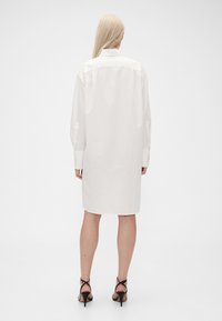 Vit skjortklänning med långa ärmar, knäppkrage, avslappnad passform, rak nederkant och detalj med bakre ok. Bärs med svarta remsandaler med klack.