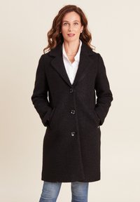 Zwarte, middellange jas van een gestructureerde wolmix met een opstaand kraagje, vijf knopen en zijzakken. Draagt men over een witte shirt.