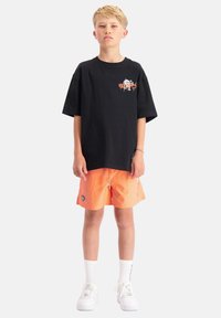 Zwarte katoenen oversized t-shirt met grafisch ontwerp op de borst, gecombineerd met oranje zwembroek met zijzakken en logo-details. Witte sneakers.