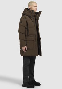 Puffjakke i mørkebrunt med hætte, med frontlynlås, store lommer og en struktureret overflade. Båret sammen med sorte jeans og støvler.