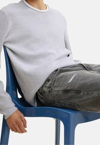 Jasnoszary sweter z teksturowanym splotem i białym kołnierzykiem, zestawiony z wystrzępionymi, ciemnoszarymi dżinsami. Osoba siedząca na niebieskim plastikowym krześle.