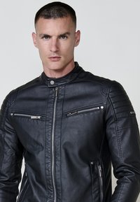 Un homme aux cheveux courts et foncés portant une veste en cuir noire avec des poches zippées sur la poitrine et les côtés, se tenant devant un fond clair et uni.