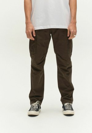 Person iført mørkebrune cargobukser, sort-hvide sneakers og en hvid skjorte med hænderne afslappet ned langs siderne.