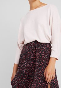 Hellrosa gestrickter Pullover mit Bootsausschnitt, kombiniert mit einem schwarzen Rock mit rot verstreuten Punkten und einem gebundenen Taillendetail.