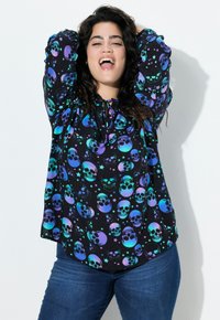 Schwarze Bluse mit buntem Schädel- und Sternmuster, mit Rundhalsausschnitt und Bindedetail. Dazu kombiniert mit blauen Jeans, die eine lockere Passform zeigen.