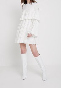 Robe blanche plissée avec des détails en volants superposés et des manches longues, associée à des bottes blanches jusqu'au genou. Texture lisse, design minimaliste.